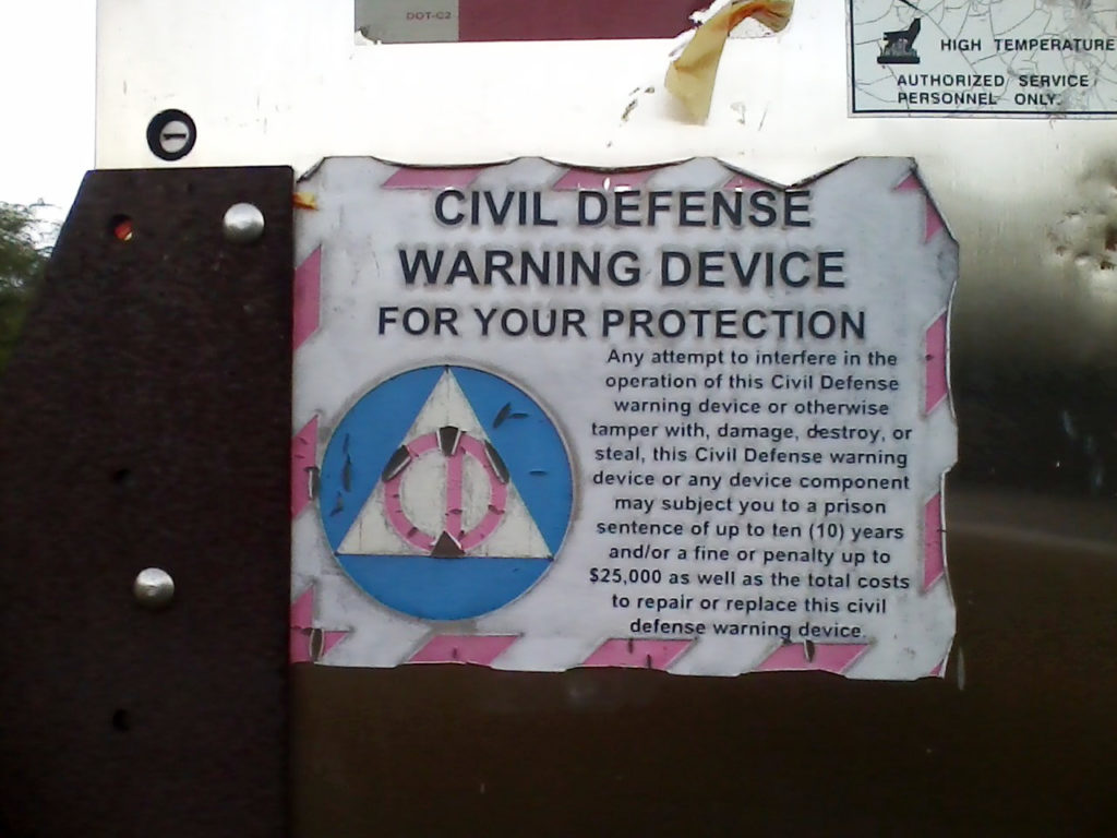 The sign from the base of a tsunami warning siren tower, which is labeled 'Civil Defense Warning Device' and uses an outdated, 1950s Civil Defense logo on it.
